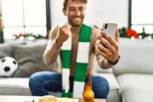 Homem vibrando com celular na mão, comida e cerveja à frente, ilustrando vício em apostas.