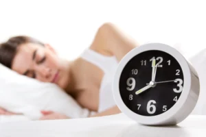 Mulher tem problemas com hornônio do sono. Ela está deitada na cama e tem um relógio à sua frente.